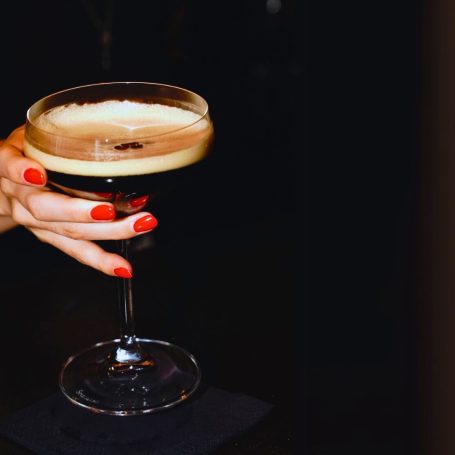 Hand mit rotem Nagellack hält ein Cocktailglas mit schaumigem Espresso-Martini.