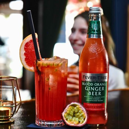 Ein Glas mit alkoholfreiem Blutorangen-Ingwerbier Cocktail und einer Limettenscheibe, im Hintergrund eine Person.